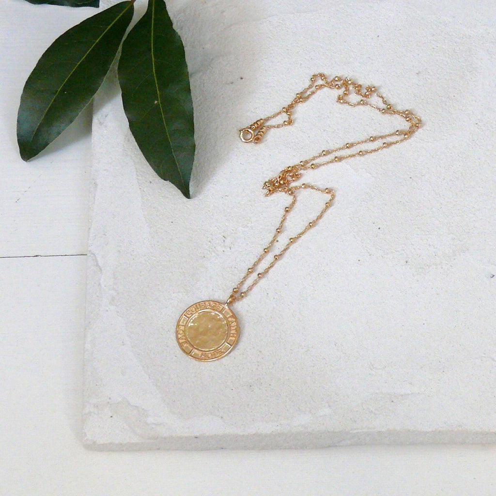 Gold pendant necklace from the Courage collection by Misia Mae on a white cement background